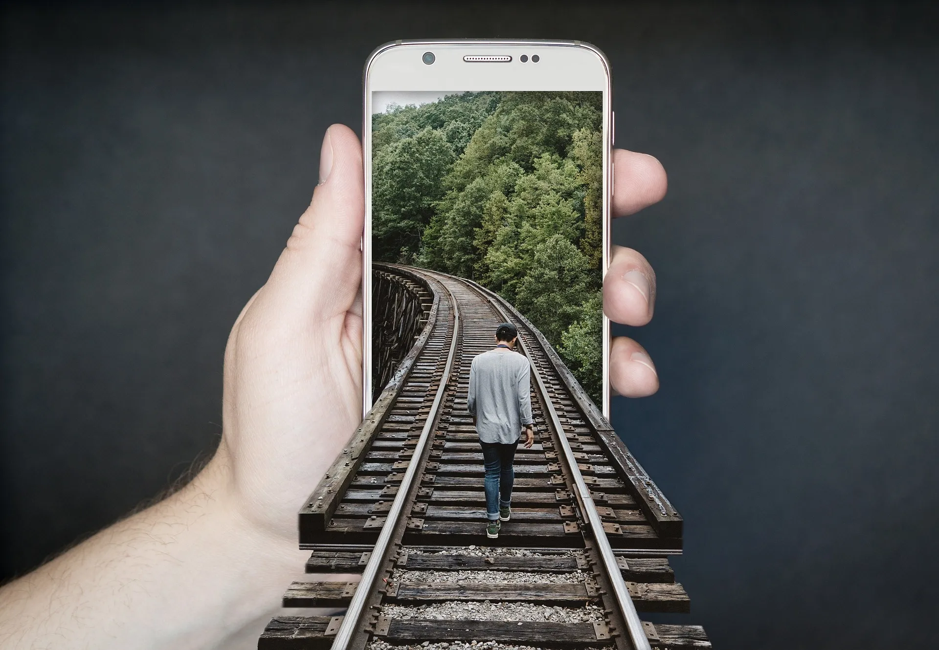 Eine Hand die ein Smartphone hält, aus dessen Bildschirm ein Gleis ragt